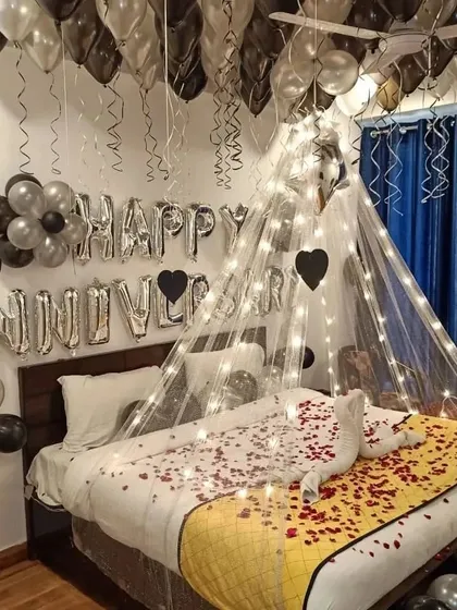 An elegant anniversary surprise using a black and silver color scheme. The setup includes a canopy with fairy lights, "Happy Anniversary" foil balloons, and a swan towel arrangement on a bed of rose petals.