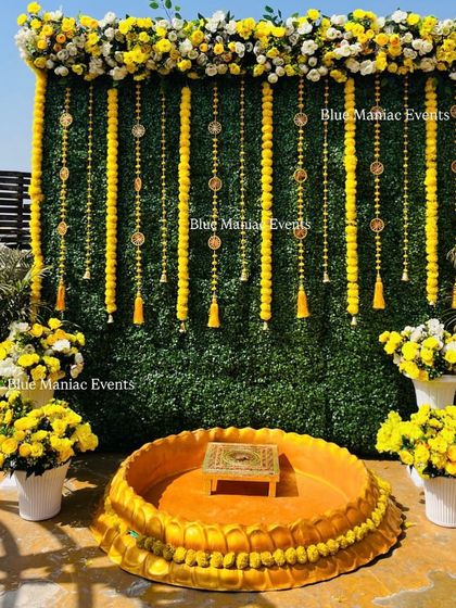 A sunny outdoor Haldi setup for the groom. The backdrop is a green wall adorned with marigold garlands, surrounded by pots of yellow flowers, creating a fresh and natural look.