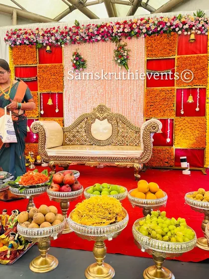 A beautiful baby shower setup with a backdrop of orange marigolds and hanging jasmine strings. The ornate golden sofa and a neat display of fruits in silver bowls complete this traditional and elegant look.