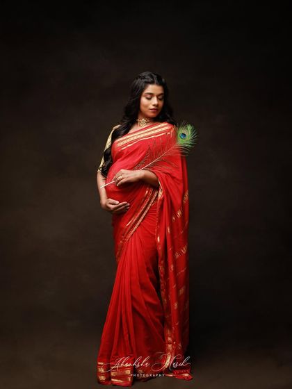 A serene and graceful portrait in a red saree, holding a peacock feather. The dark, moody background makes the vibrant color of the saree stand out beautifully.
