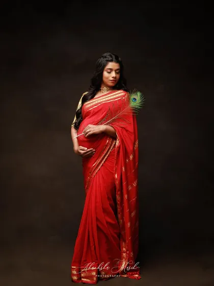 A serene and graceful portrait in a red saree, holding a peacock feather. The dark, moody background makes the vibrant color of the saree stand out beautifully.