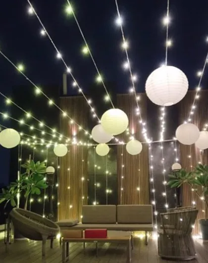 A beautiful terrace setup for a New Year's party, perfect for any evening celebration. The space is decorated with strings of fairy lights and hanging white paper lanterns, creating a warm and inviting atmosphere.