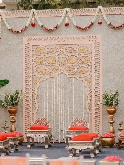 We blend tradition with artistry in our designs. This engagement stage features an intricately carved pink jharokha-style backdrop, elegant silver seating with red cushions, and traditional brass lamps, all framed by banana plants for an authentic South Indian touch.