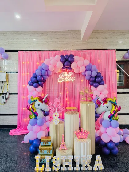 A magical unicorn theme birthday party with a full decorative arch. The setup features a pink curtain backdrop with fairy lights, a pink and purple balloon arch, large unicorn foil balloons, and various props like a crown and the Eiffel tower.