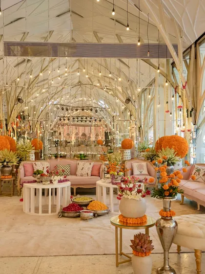 A beautiful indoor Mehendi setup, transforming the space with pastel lounges, marigold accents, and traditional garlands for a "pooja to party" vibe.