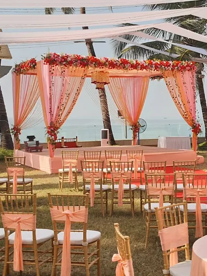 A beautiful peach-themed wedding ceremony on a lawn overlooking the ocean. The decor is light and elegant, with matching chair ties and a floral mandap that perfectly complements the seaside setting.