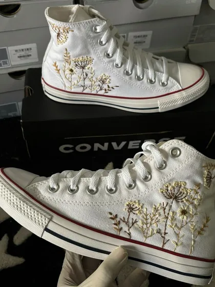 A pair of classic white Converse All-Stars with delicate, custom floral embroidery in gold and brown thread.