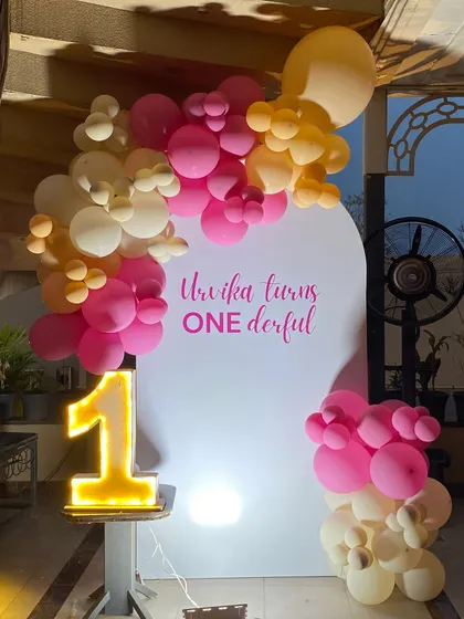 A simple and modern 'ONEderful' first birthday setup. The clean white backdrop is accented with a pink and orange balloon cluster and a bright, light-up number '1'.