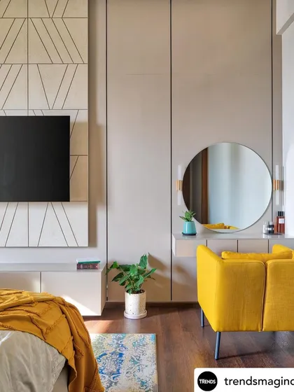 A bedroom from a feature in Trends Magazine. The design uses a neutral color palette with a pop of mustard yellow, a geometric pattern behind the TV, and a simple, elegant vanity area.