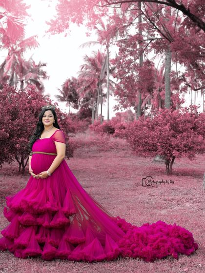 A stunning outdoor portrait with a pink theme. The mother-to-be wears a ruffled gown and crown, surrounded by pink-toned foliage.