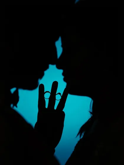 A creative silhouette of the couple, their hands forming a 'V' to frame their wedding rings against a bright blue background. A unique and artistic engagement shot.