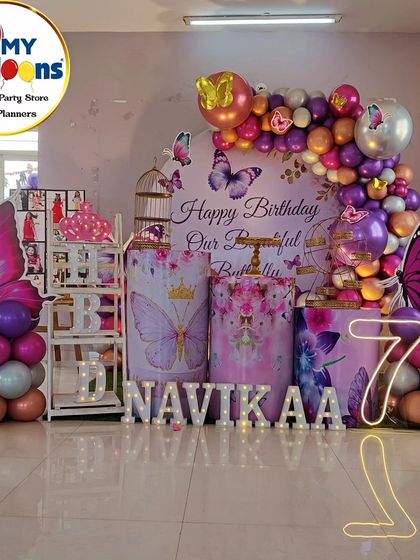 A full-stage butterfly-themed seventh birthday. The decor includes a photo collage, a 'Happy Birthday Our Beautiful Butterfly' backdrop, and a neon '7'.