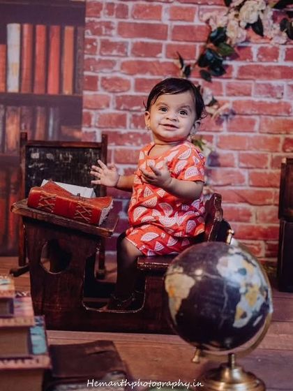 A baby boy sits in a desk-like prop in a library-themed setup, complete with a globe and books. This is an example of a creative 'sitter session' for babies who can sit up.