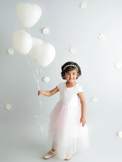 A little girl in a beautiful white dress holds a bunch of heart-shaped balloons. Props like balloons can add a touch of whimsy and joy to any child's portrait session.