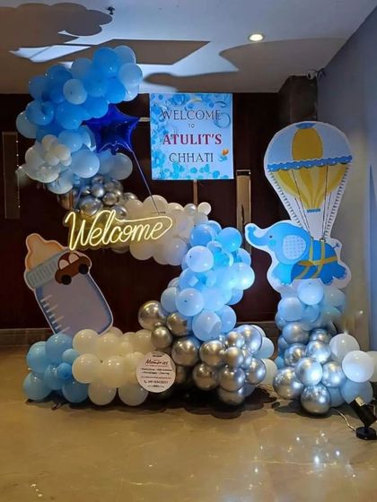 A multi-prop setup for a baby's Chhathi ceremony. This decoration includes a "Welcome" neon sign, balloon clouds, and themed cutouts like a hot air balloon and a baby bottle, all in a cohesive blue and white theme.