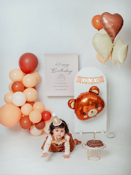 This little one is crawling towards her first birthday cake in a studio filled with warm, earthy-toned balloons and teddy bear decorations.