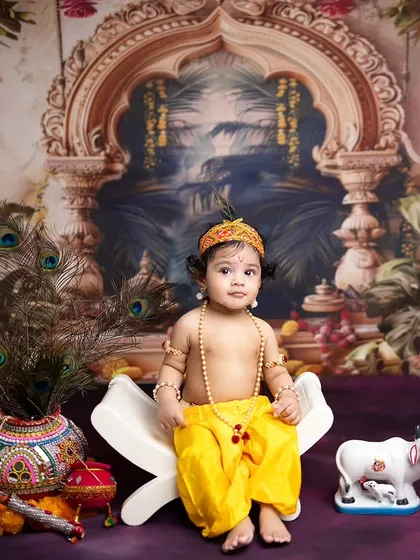 A full portrait of the baby dressed as Lord Krishna, seated on a prop against a beautiful, traditional Indian backdrop.