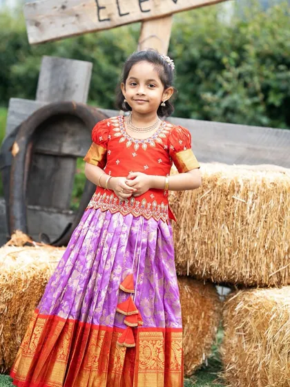 Our little client looks absolutely radiant in her custom Kanchi Pattu lehenga. The bright red peplum blouse has detailed embroidery, perfectly complementing the lavender silk skirt with its wide, golden zari border.