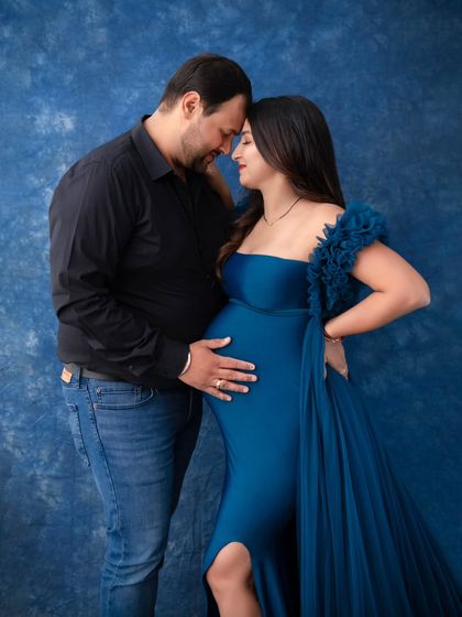 An intimate and powerful portrait against a deep blue backdrop. The couple's foreheads are touching as they both gently hold the baby bump, a symbol of their unity and shared love.