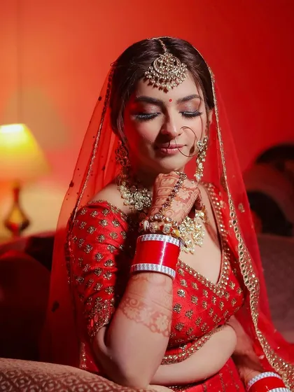 A bride in a classic red lehenga, seated on a couch, with warm, dramatic lighting.