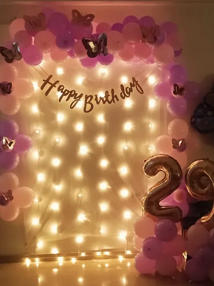 A sweet and simple butterfly-themed decoration for a 29th birthday. A fairy light curtain is framed by a pink and purple balloon arch.