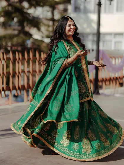 A joyful bride-to-be in a vibrant green and gold outfit twirls happily during her outdoor Mehandi ceremony.