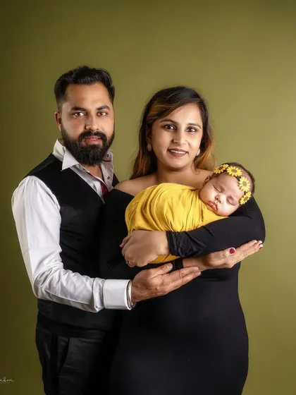 A classic family portrait against a warm, olive green backdrop. The parents hold their newborn daughter together, creating a timeless image of unity and love.