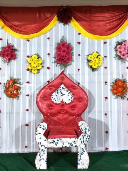 A colorful backdrop for a half saree decoration at home, featuring bright red and yellow drapes. The setup includes a unique red throne chair and assorted flower arrangements.