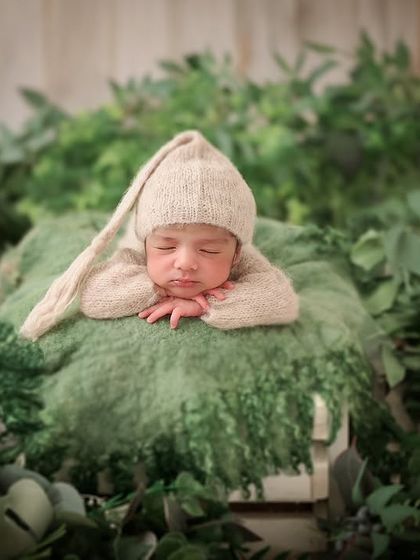 Surrounded by lush greenery, a newborn in a beige pixie hat sleeps on a soft green cushion, creating a peaceful, nature-filled scene.