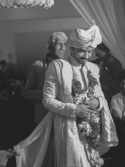 A candid black and white shot of the groom during the wedding ceremony, with a proud family member in the background.