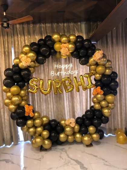 A bold and beautiful birthday decoration with a black and gold balloon ring. The setup is personalized with foil letters spelling out the name "Surbhi".