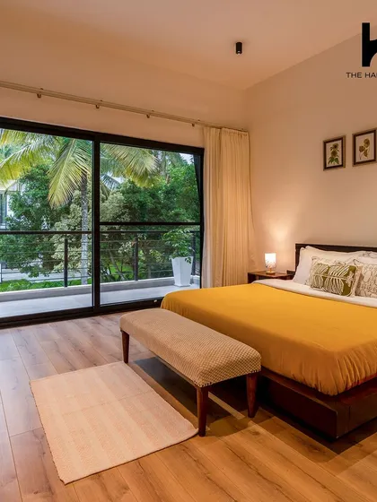 A beautifully designed bedroom with a view. The room features a comfortable bed with a pop of yellow, hardwood flooring, and a large sliding glass door that opens onto a balcony overlooking the lush greenery.