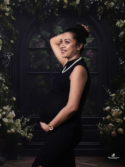 A chic and happy solo portrait. The mom-to-be, in a sleek black dress, smiles brightly against a dark, moody backdrop framed with white flowers.