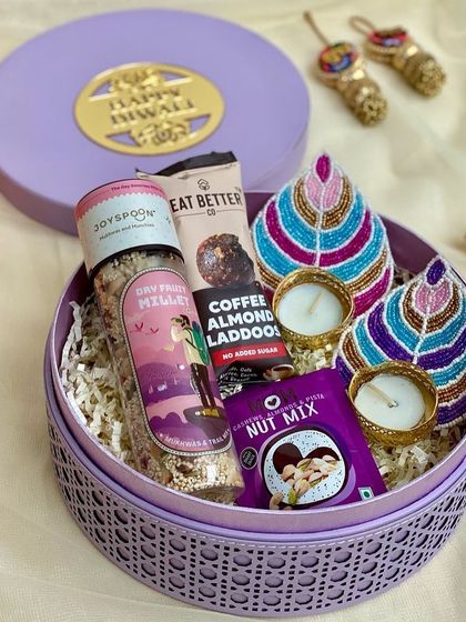 A round rattan box in a lovely lilac color, filled with healthy treats and hand-painted diyas for a unique Diwali gift.
