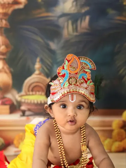 A charming close-up of baby Krishna making a cute face, highlighting the traditional tilak and ornate crown.