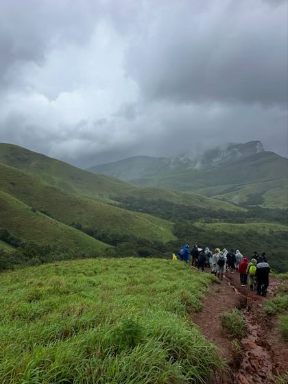 A line of trekkers disappears into the vast green landscape of Kudremukha under dramatic monsoon clouds.