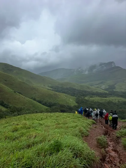 A line of trekkers disappears into the vast green landscape of Kudremukha under dramatic monsoon clouds.