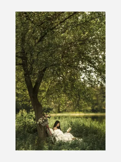 A portrait of solitude and peace. The mother-to-be reads quietly under a large, shady tree by the water, a serene and beautifully composed image from a styled session.