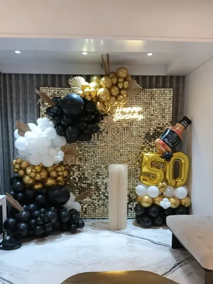 A stylish 50th birthday decoration for a home party. It features a gold sequin wall, a black and gold balloon arrangement, and a large whiskey bottle foil balloon for a personal touch.