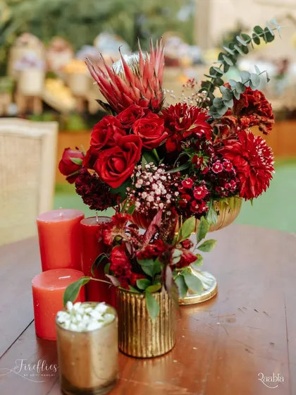 A lush floral centerpiece combining red roses, dahlias, and protea in a brass vase. The arrangement's deep red tones and varied textures perfectly captured the 'Laal Ishq' theme.