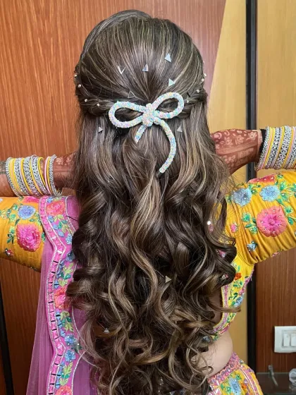 A playful and pretty half-up hairstyle with soft curls, decorated with a sparkling bow and small pearl details. Perfect for a fun mehendi or sangeet look.
