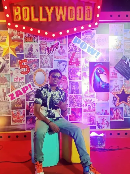 Posing in front of a Bollywood-themed backdrop. I am your go-to DJ for desi parties, with an extensive library of Bollywood and regional hits.