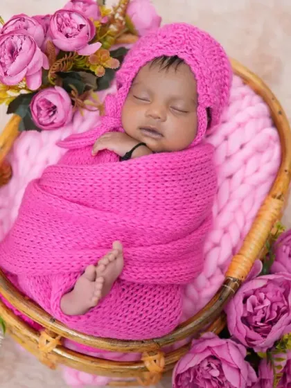A newborn is wrapped in a bright pink knit swaddle and matching bonnet, nestled in a basket surrounded by flowers. This cozy and colorful setup is perfect for a sweet and classic baby portrait.