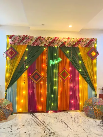 A colorful backdrop for a Mehndi ceremony, using drapes in shades of yellow, green, and pink, accented with fairy lights and a 'Mehndi' neon sign.