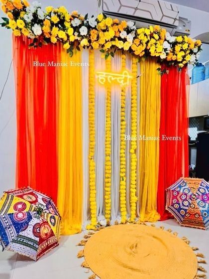 A simple and sweet Haldi backdrop with red, orange, and yellow drapes, marigold garlands, and traditional umbrellas.