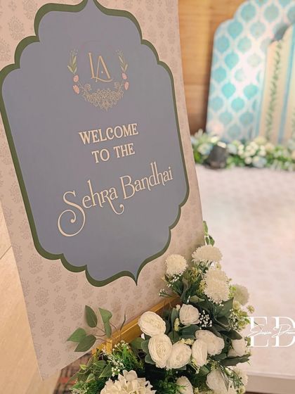 An elegant welcome sign for a 'Sehrabandi' ceremony, featuring a custom monogram and beautiful calligraphy. The sign is adorned with a delicate arrangement of white flowers.