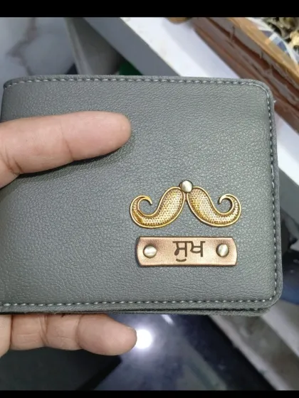 A close-up of a grey personalized wallet with a mustache charm and a name engraved in Hindi script, held in hand to show its texture and size.