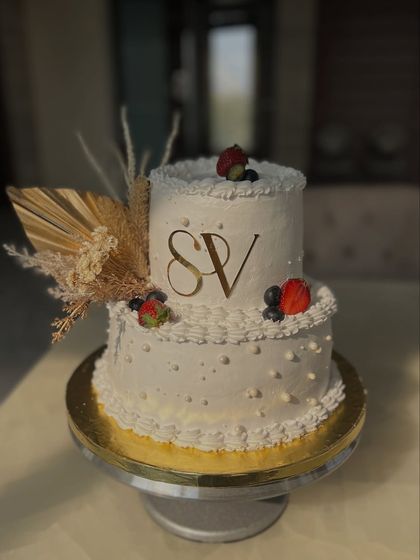 A classic two-tiered engagement cake with the couple's initials. The design is kept clean and elegant with dried pampas grass, fresh berries, and simple piping.