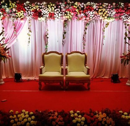 A classic and elegant stage for a formal ceremony. This setup features two ornate chairs for the guests of honor, set against a backdrop of soft pink drapes and a lush floral border.