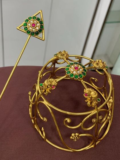 This exclusive gold-plated designer bun cover is a stunning hair accessory for weddings and special occasions. The intricate frame is adorned with floral motifs set with green and red stones.